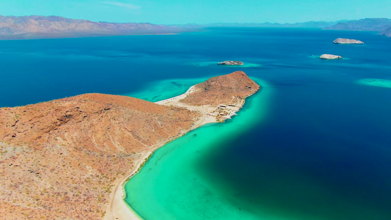 Aerial frame over Playa Santispac showing a rocky desert peninsula, pale sand, turquoise shallows, distant islands, hazy mountains, and no visible people.