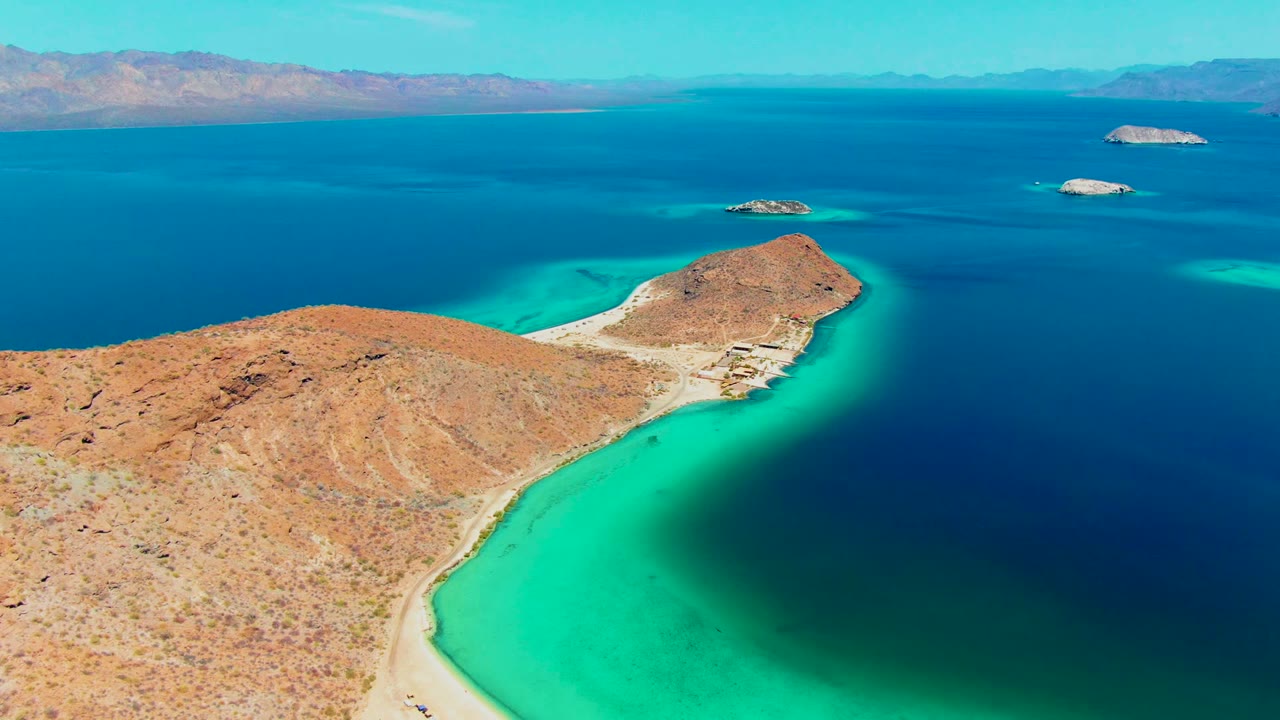 Aerial frame over Playa Santispac showing orange-brown desert terrain in the foreground, a bright turquoise bay edge, distant islands, and no visible people.