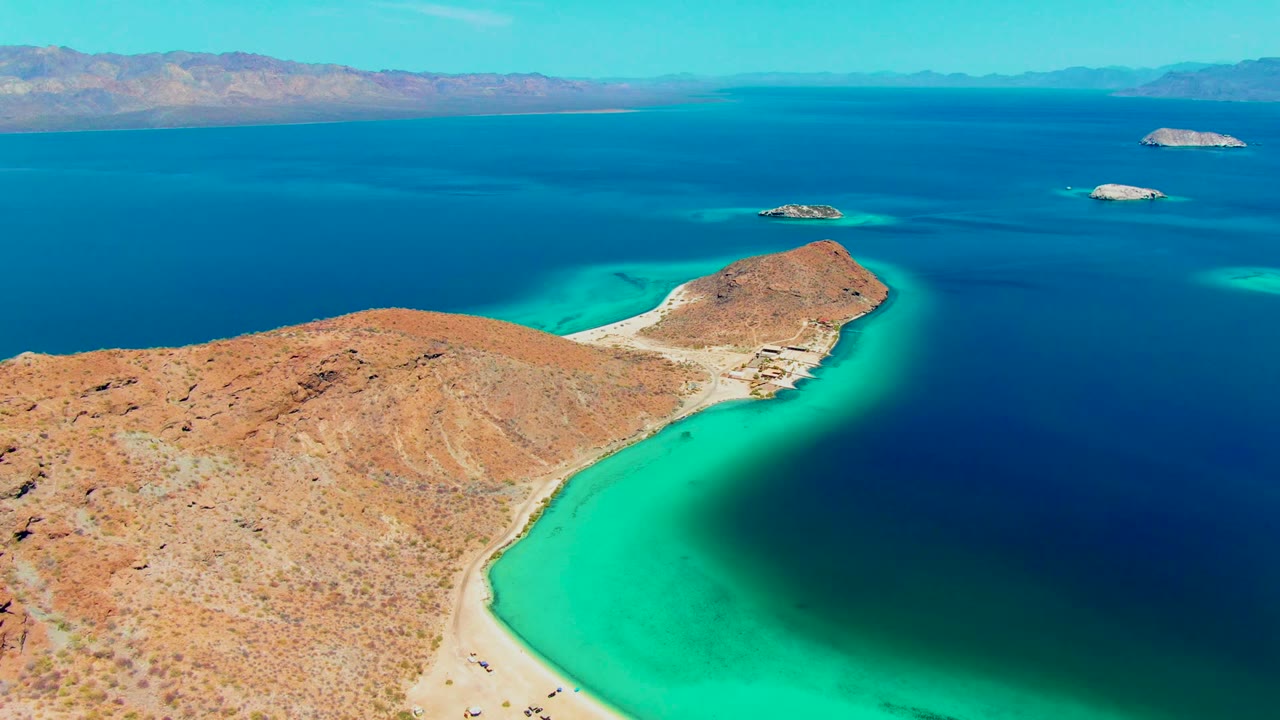 At 0:00, Playa Santispac appears from above with a dry rocky peninsula in the foreground, a curved sandy beach, bright turquoise shallows, dark blue open water, small offshore islands, distant mountains, and tiny beachgoers visible.