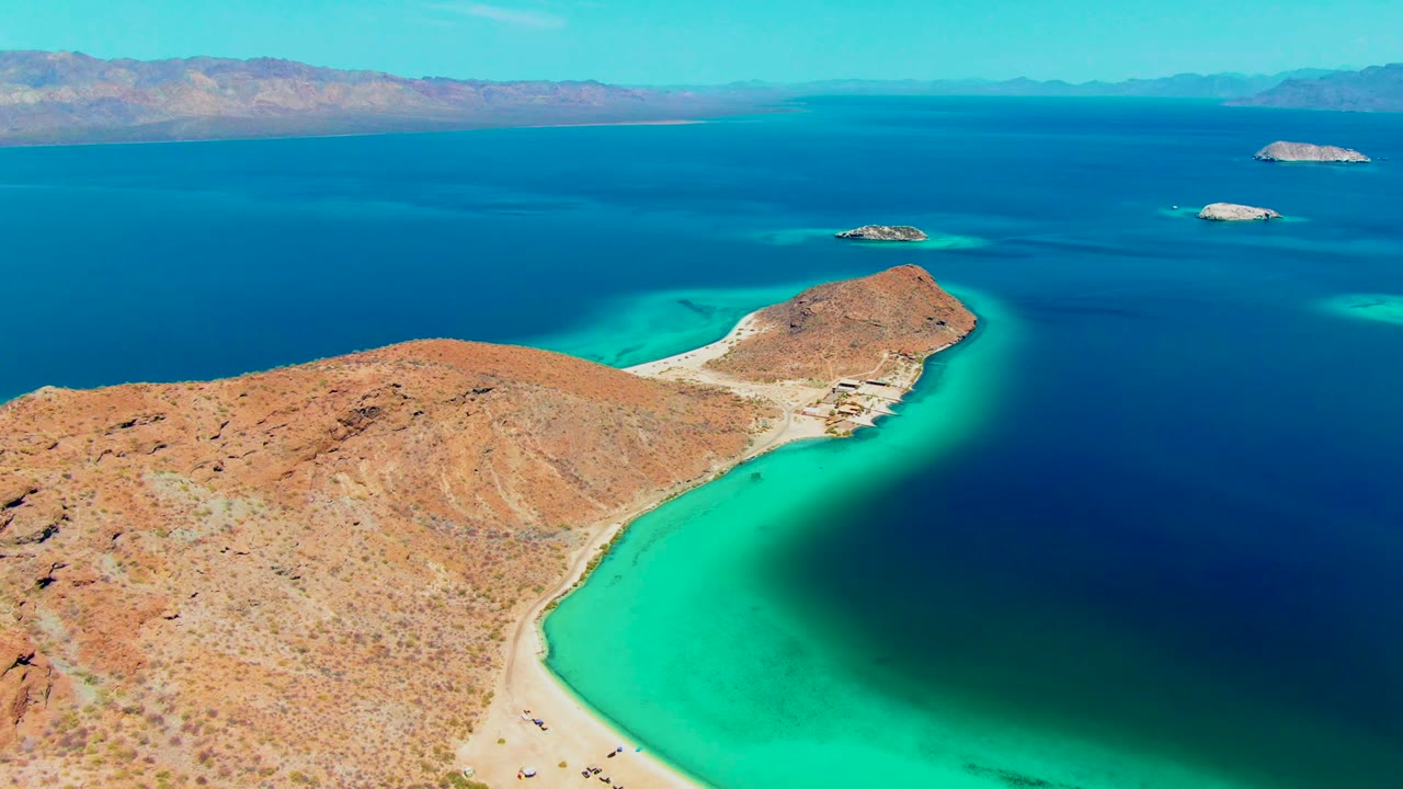 Aerial coastal view with arid peninsula in front, turquoise bay, dark blue sea, small islands, and mountain horizon.