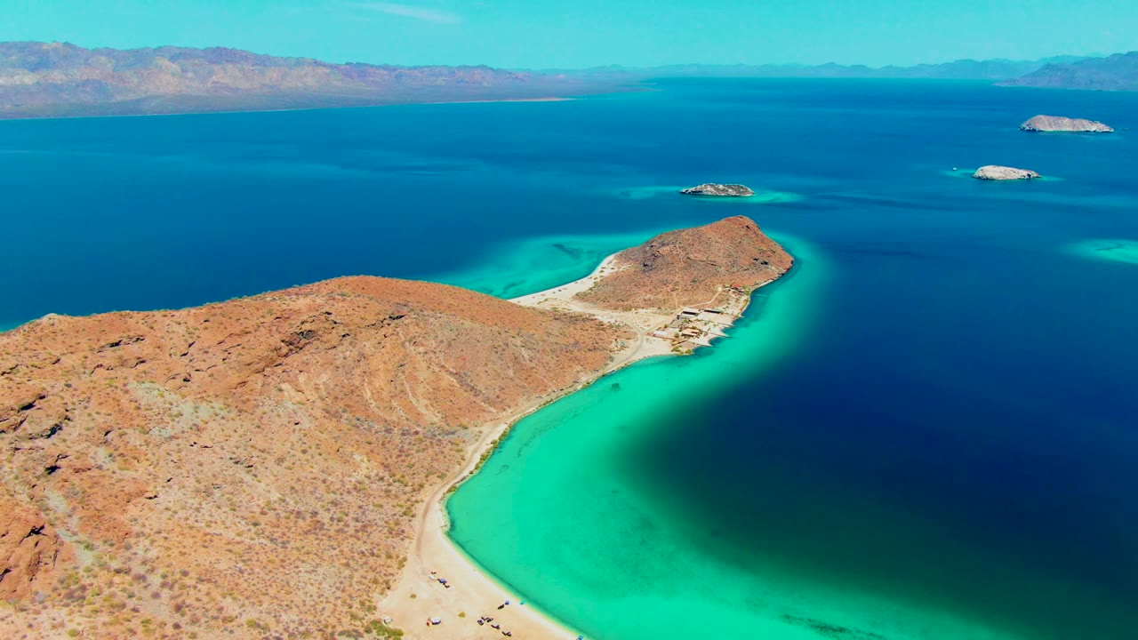 At 0:02, Playa Santispac remains in a wide drone composition, with scrub-covered desert terrain in front, a narrow sandy shoreline below, green-blue shallows at the bay edge, and small islands beyond the headland.