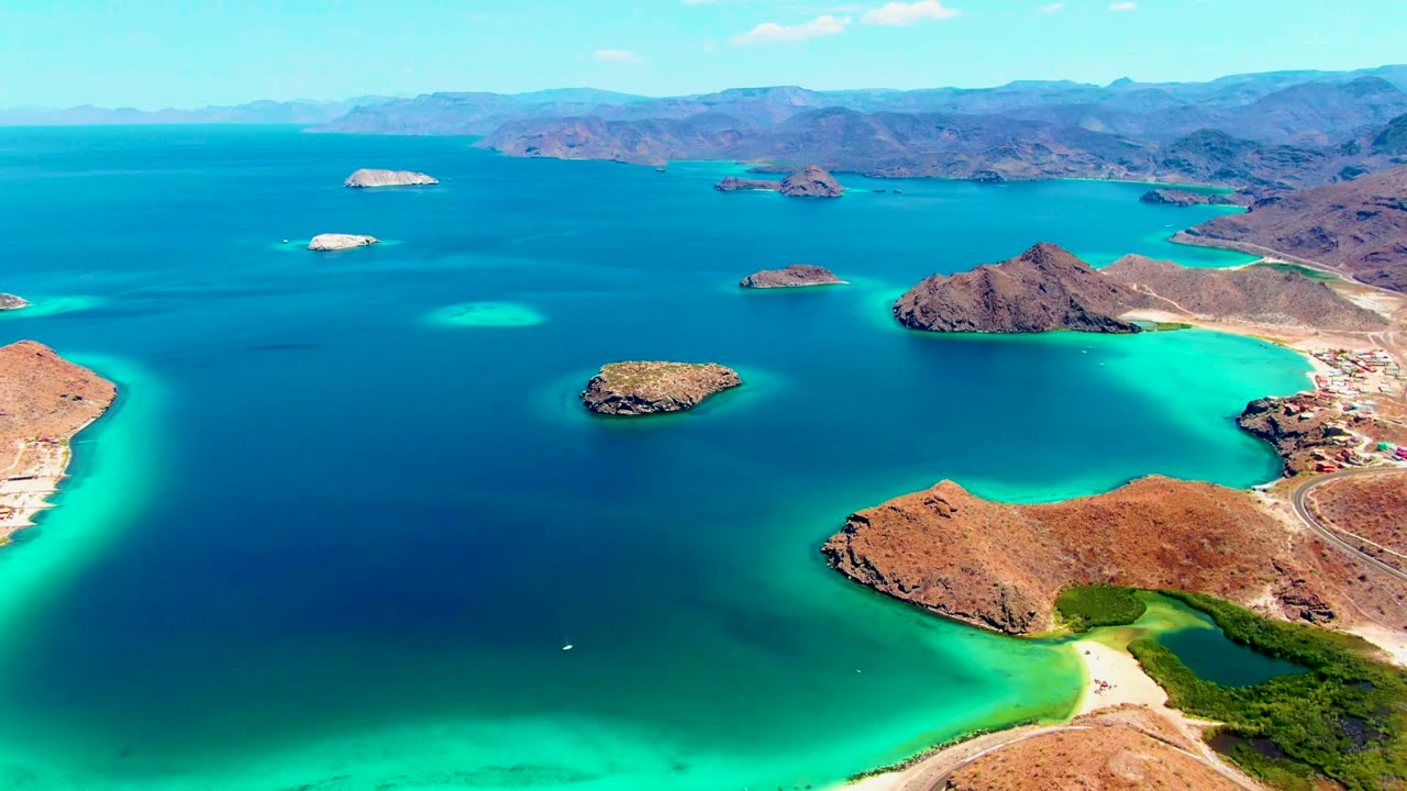 At 0:00, an aerial view over Playa Santispac shows a small white boat low in the bay, turquoise shallows near the beach, rocky islands across deep blue water, distant mountains, and no visible people.