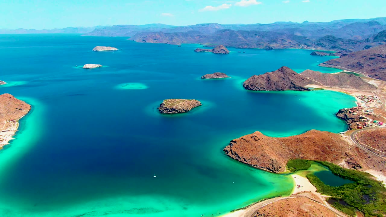 Small boat in blue water with turquoise shallows, rocky islands, and mountains beyond.