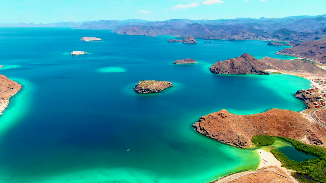 A wide aerial frame over Playa Santispac shows turquoise shallows, deep blue water, rocky islands, tan desert hills, a sandy beach, and no visible people.