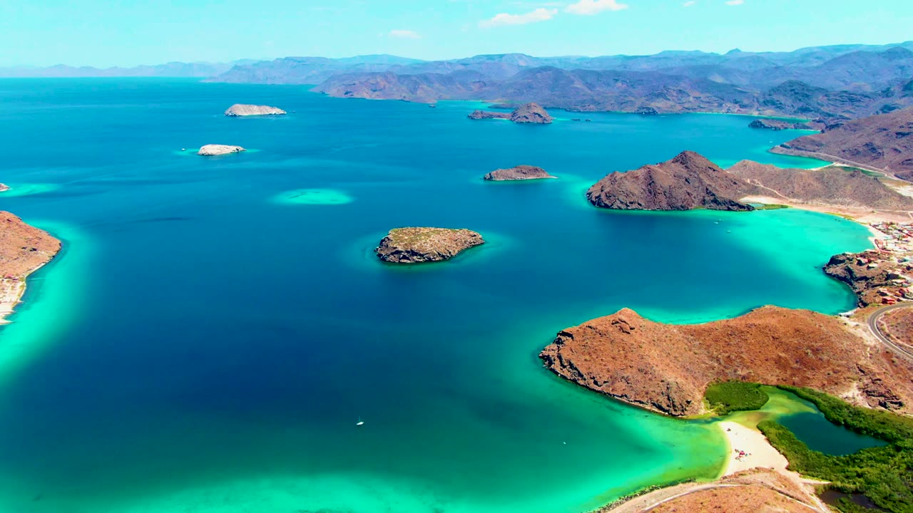 Wide drone view of blue ocean, small rocky islands, turquoise shallows, and arid coastline.