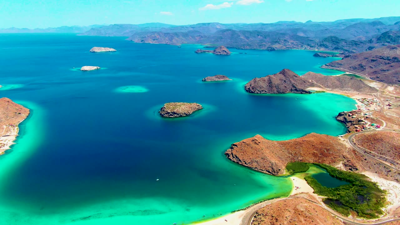 Aerial view over Playa Santispac with rocky islands in deep blue water, bright turquoise shallows along the coast, desert mountains in the distance, and no visible people.