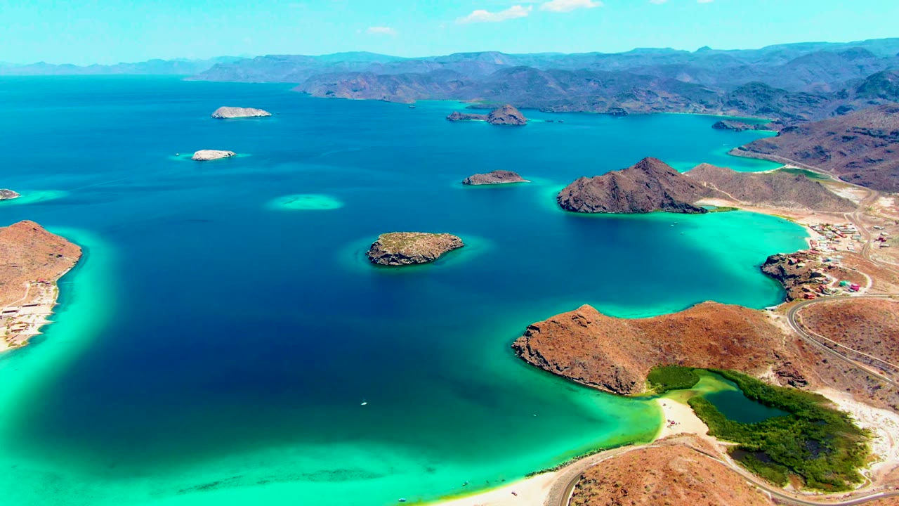 Aerial frame over Playa Santispac with deep blue bay water, rocky islets, vivid shallow water near the desert shore, mountain ridges beyond, and no visible people.