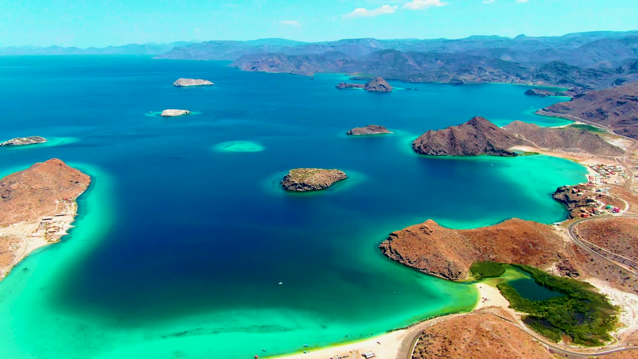 Opening aerial frame over Playa Santispac, Baja California Sur, showing turquoise shallows, deep blue bay water, rocky islands, sandy shoreline, distant mountains, and no visible people.