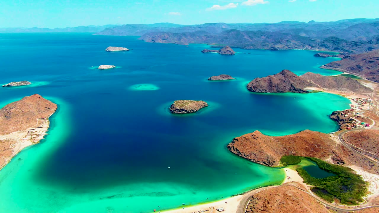 High aerial view of blue bay water, tan peninsulas, rocky islands, and pale beaches.