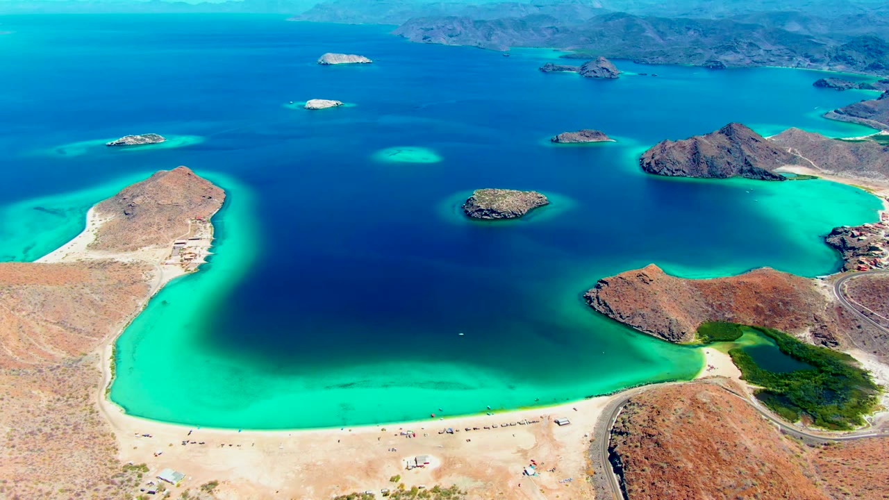 Opening aerial frame over Playa Santispac, Mexico, showing turquoise shallows around sandy beach, reddish rocky landforms, small islands, distant mountains, and no visible people.