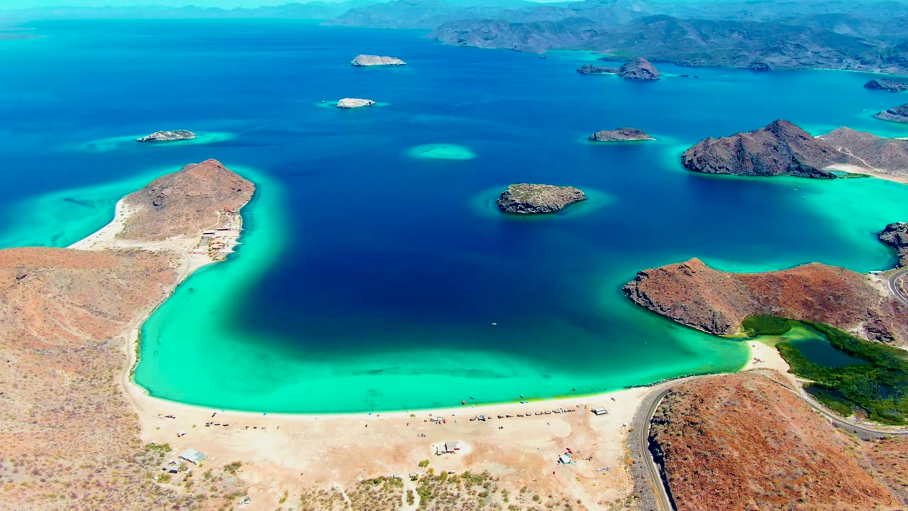Aerial view over Playa Santispac with turquoise shallows along a sandy beach, dark blue lagoon water, rocky islands, distant mountains, and no visible people.