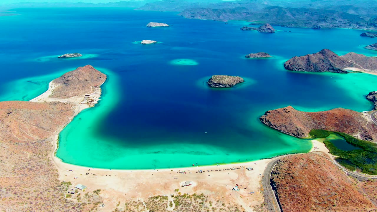 Aerial view of Playa Santispac with bright shallow water near the beach, darker lagoon water offshore, small rocky islands across the bay, desert terrain, and no visible people.