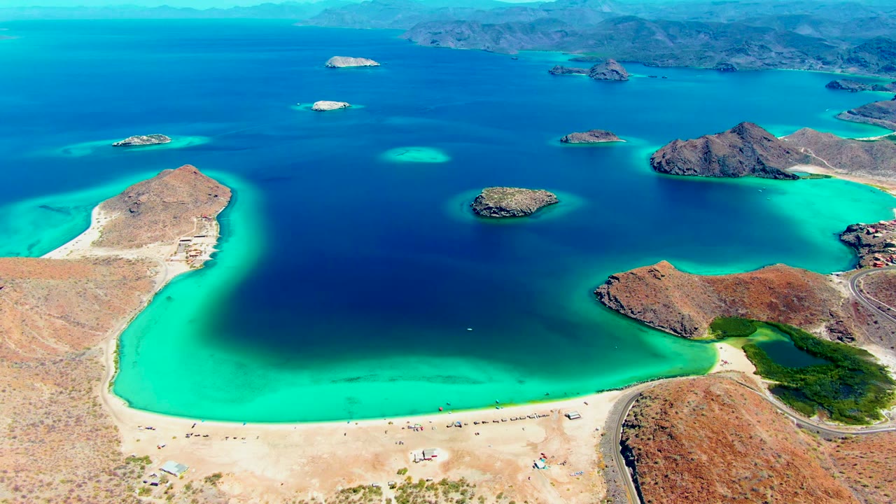 Opening aerial frame over Playa Santispac, with bright turquoise shallows curving around a sandy beach, small visible people along the shore, rocky islands offshore, and dry hills beyond.