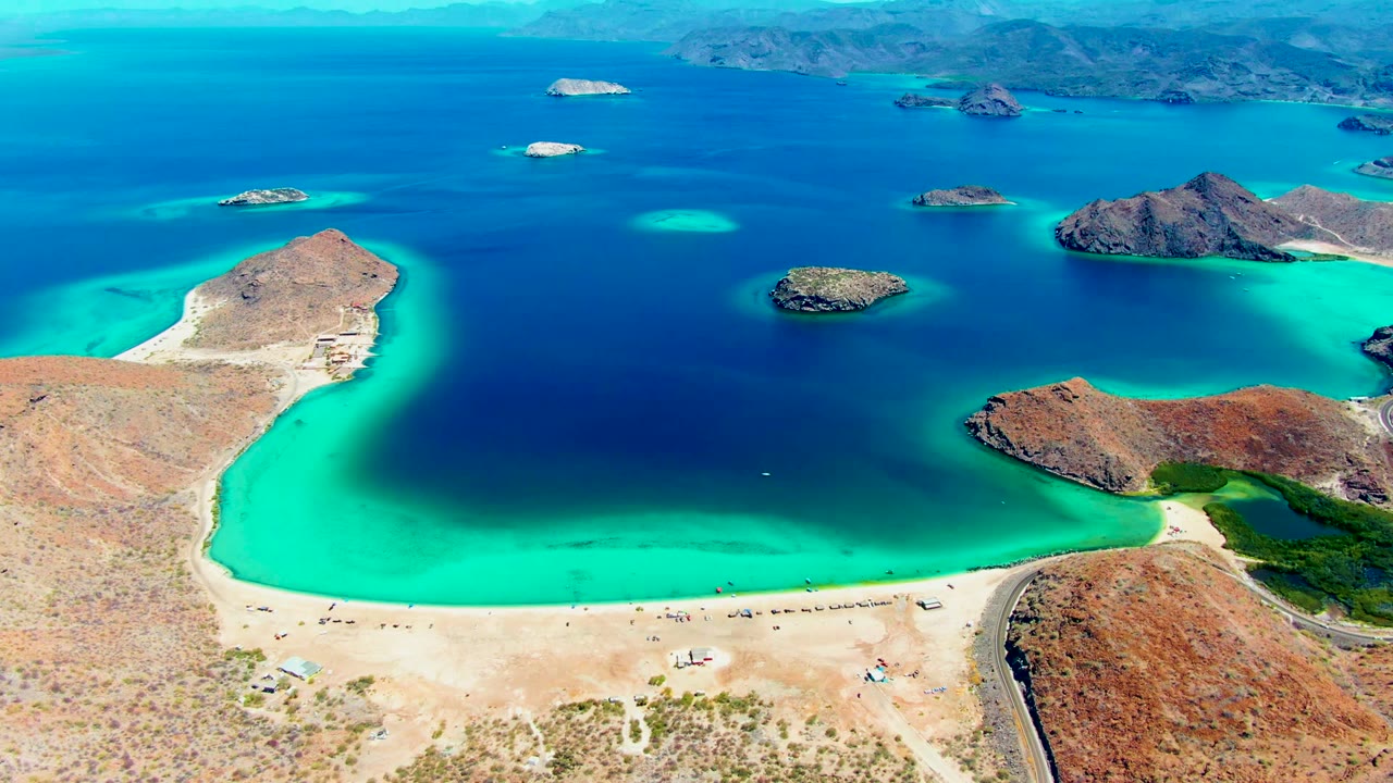 Aerial frame of Playa Santispac with a curved sandy beach in the foreground, turquoise shallows, dark blue bay water, rocky islands offshore, and tiny people and vehicles on the shore.