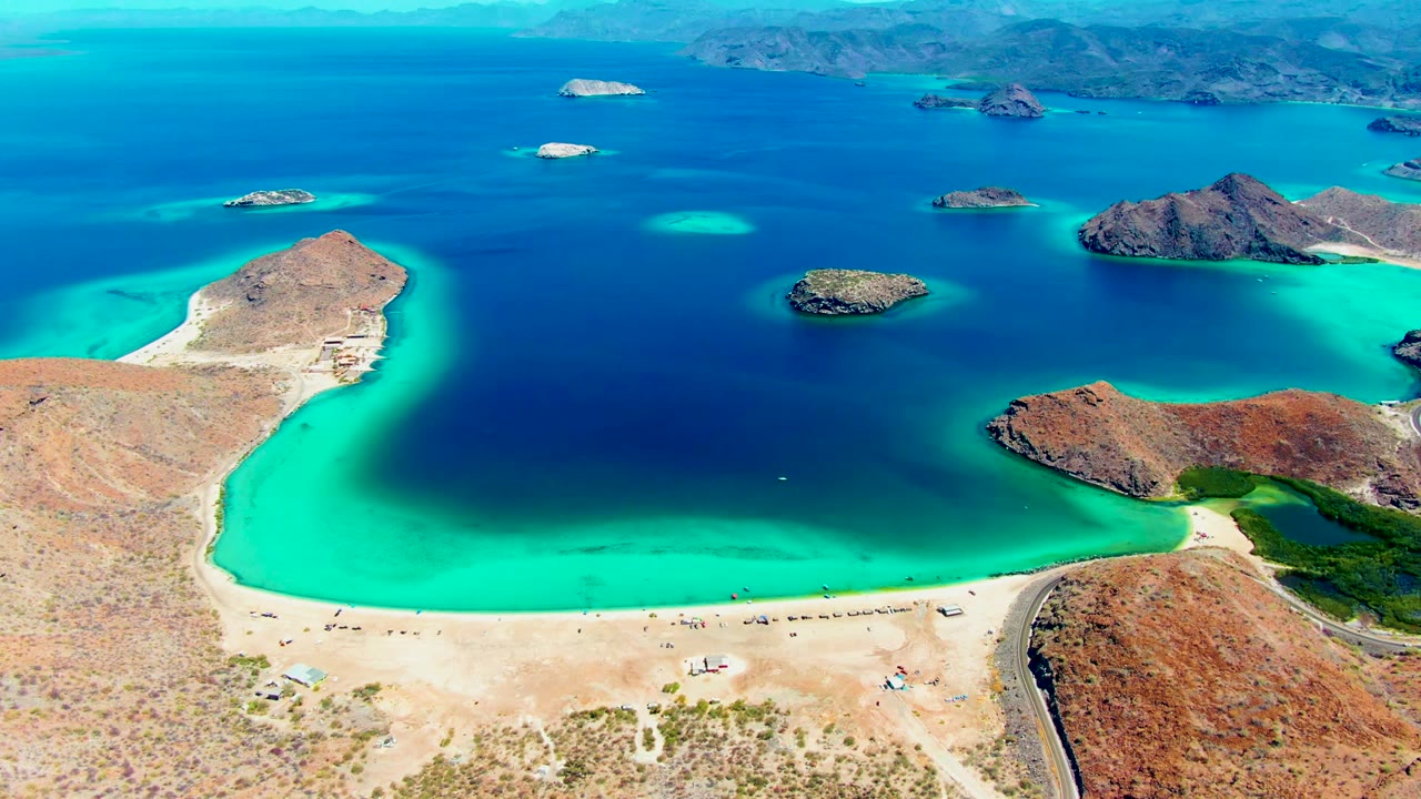 Aerial frame of Playa Santispac with the beach stretching across the lower frame, bright turquoise shallows near shore, scattered rocky islands in the bay, and dry mountains in the distance.