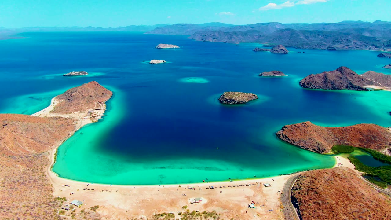 Wide aerial frame over Playa Santispac showing a pale sandy beach below, turquoise shallows, deep blue bay water, rocky islands, distant mountains, bright sky, and no visible people.