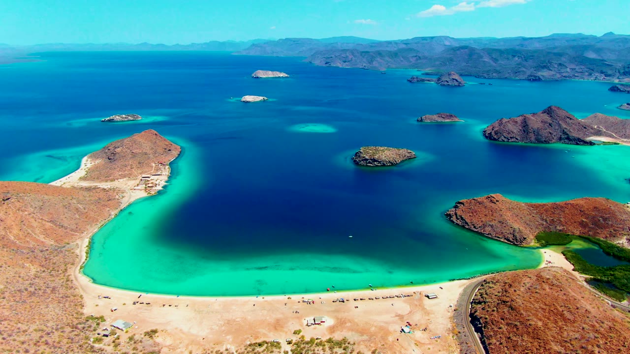Playa Santispac aerial frame showing the sandy foreground shore, a broad turquoise band along the bay edge, darker blue open water, scattered rocky islands, hazy mountain ridges, and no visible people.