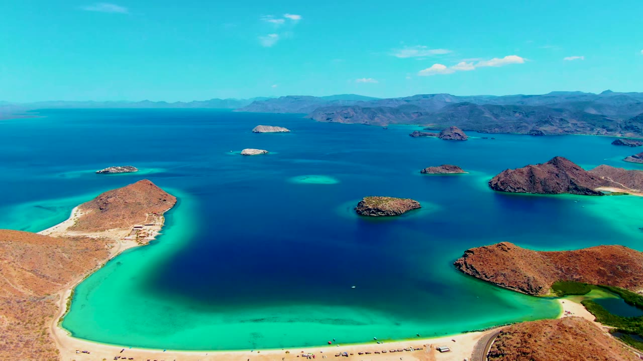 Aerial frame over Playa Santispac shows a sandy shoreline at the lower edge, turquoise shallows, several rocky islands, deep blue water, distant mountains, and no visible people.