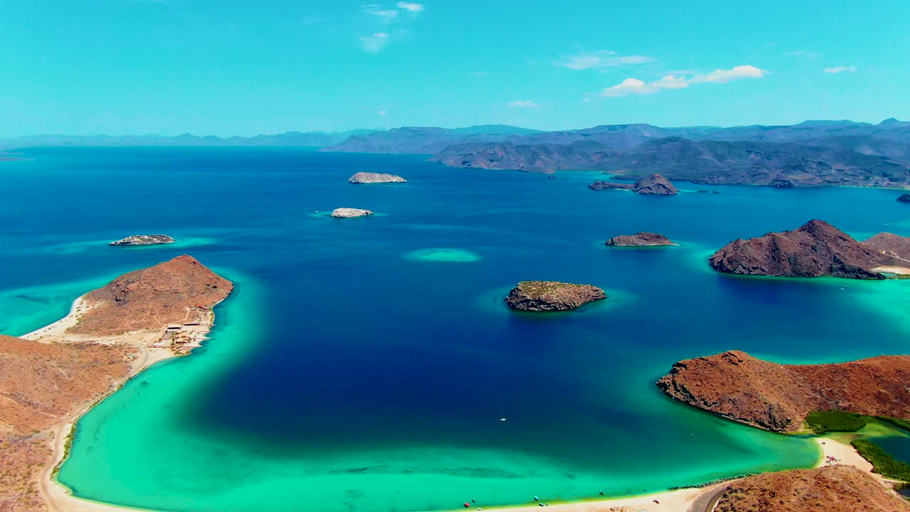 Aerial frame over Playa Santispac shows the sandy shoreline sliding toward the lower left edge, with turquoise shallows, scattered rocky islets, a broad blue bay, mountain ridges, and no visible people.