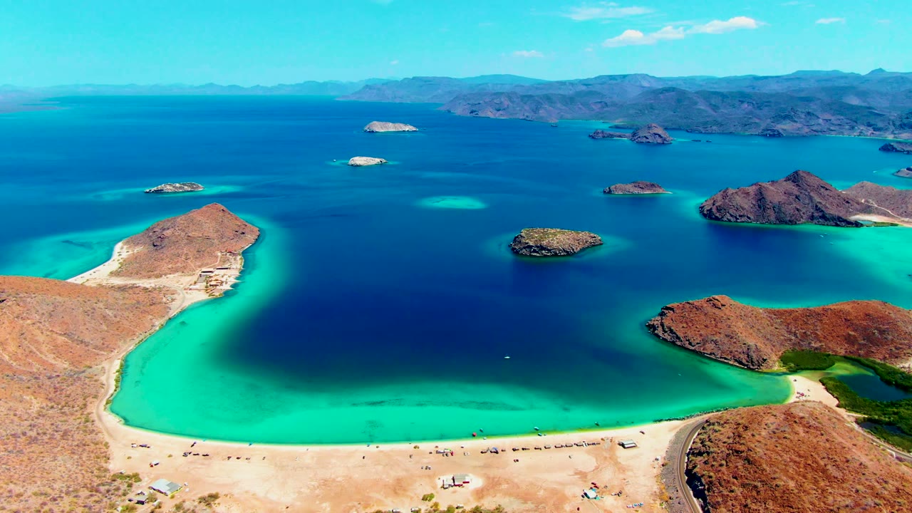 Wide drone view of turquoise shallows around a sandy beach and rocky islands.