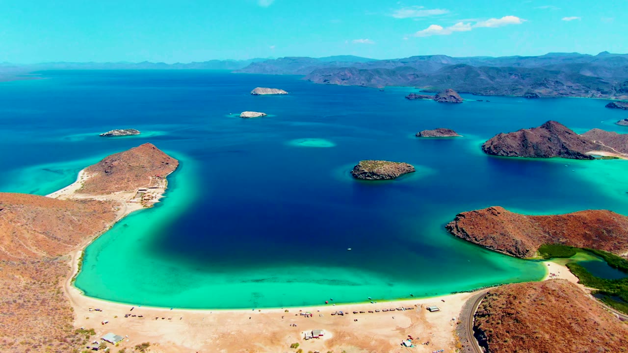 At 0:02, the Playa Santispac aerial view holds on the curved sandbar-like beach, green-blue shallows, dark open water, scattered rocky islets, arid hills, and small beachgoers and vehicles below.