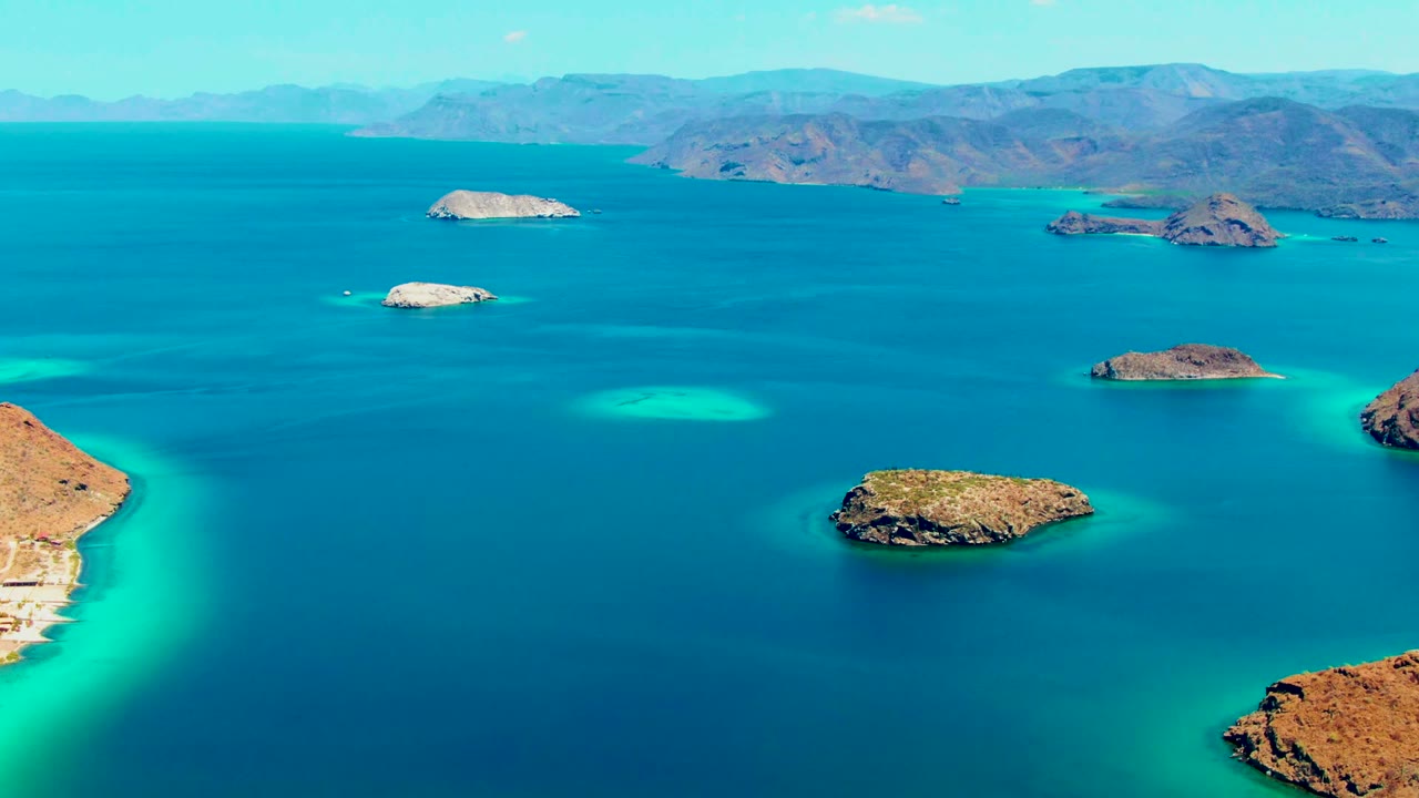 Aerial frame over Playa Santispac showing small rocky islands in turquoise shallows and deeper blue water, with distant mountains and no visible people.