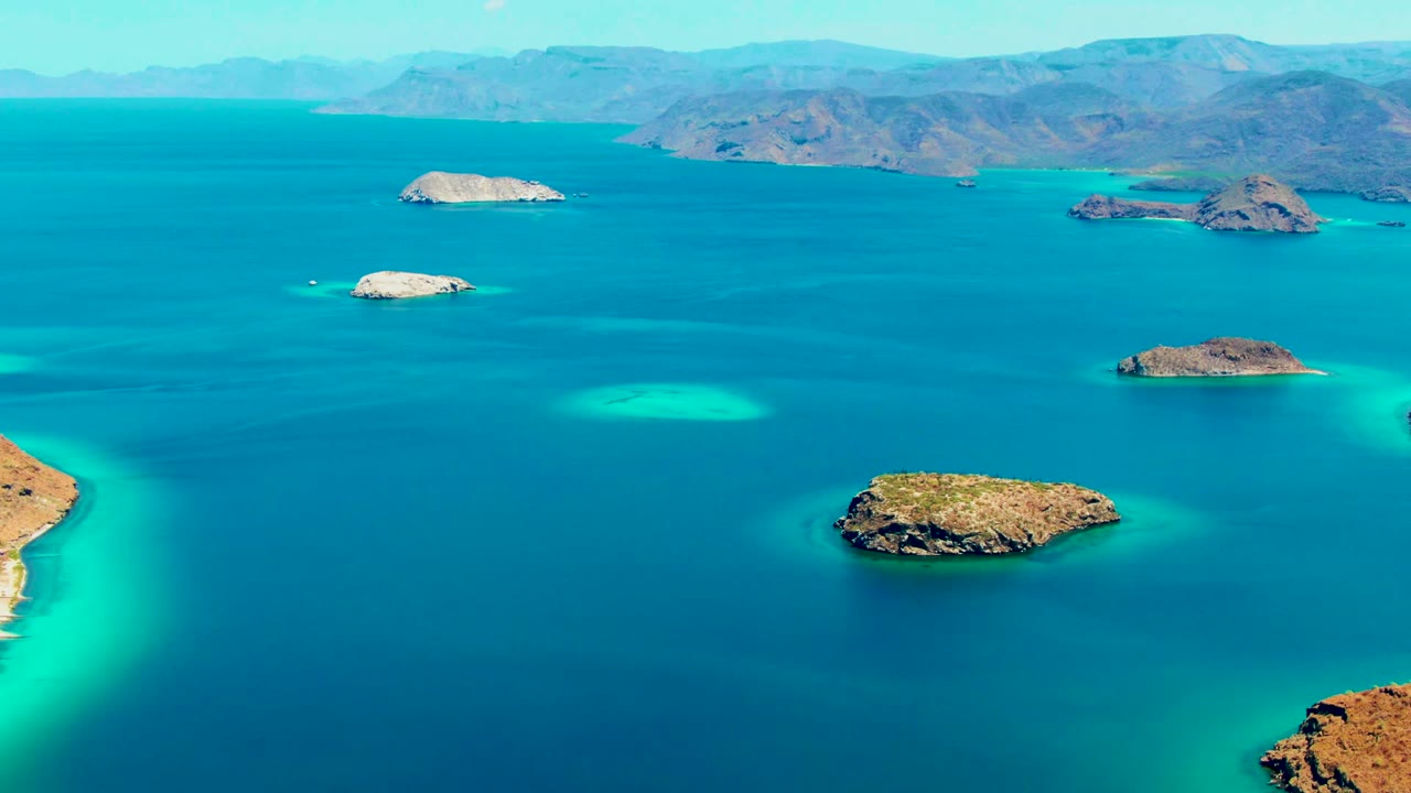 Aerial frame from the slow pan showing scattered rocky islands across Playa Santispac waters, a broad blue channel, turquoise shoals, and no visible people.