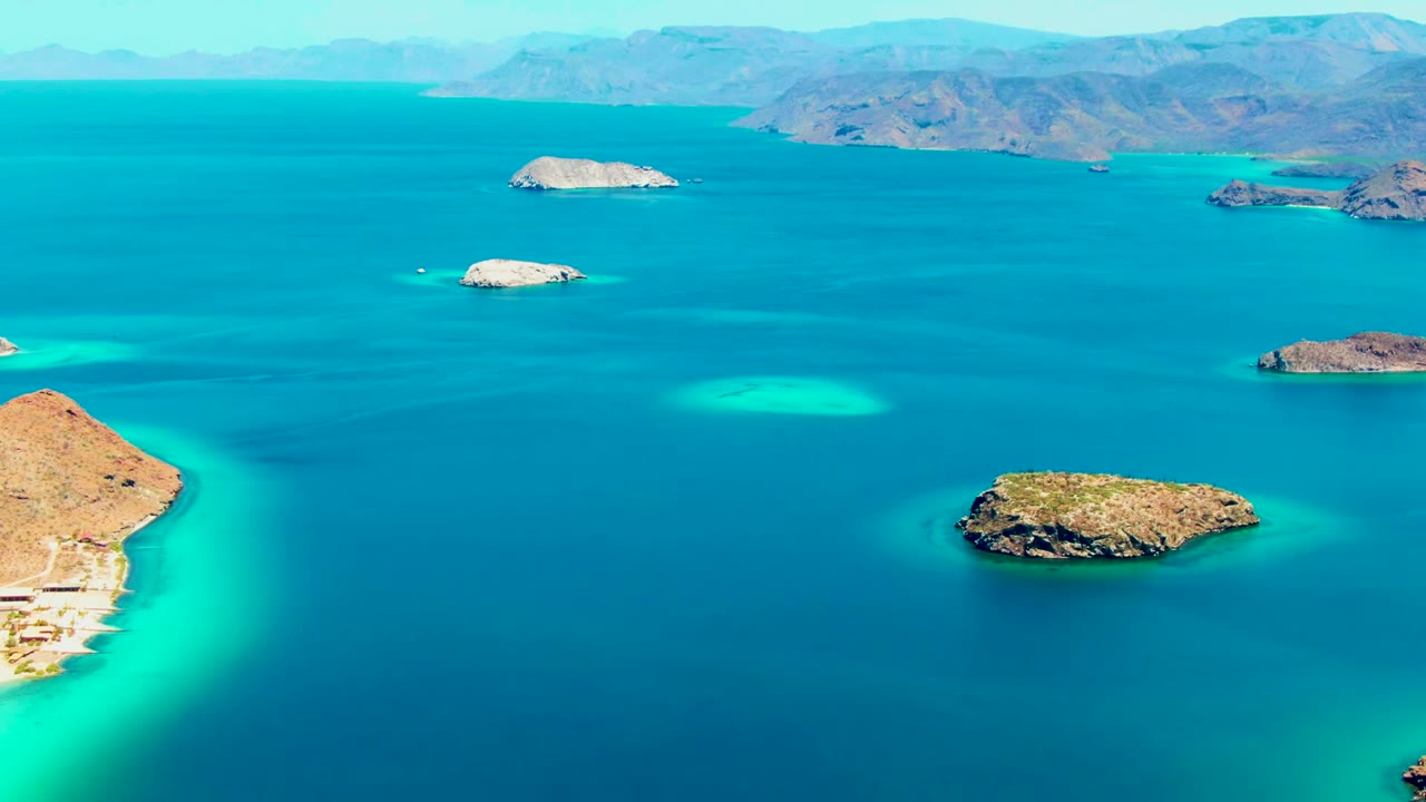 Opening aerial frame over Playa Santispac, Baja California Sur, showing rocky islands in blue and turquoise water with distant mountains and no visible people.
