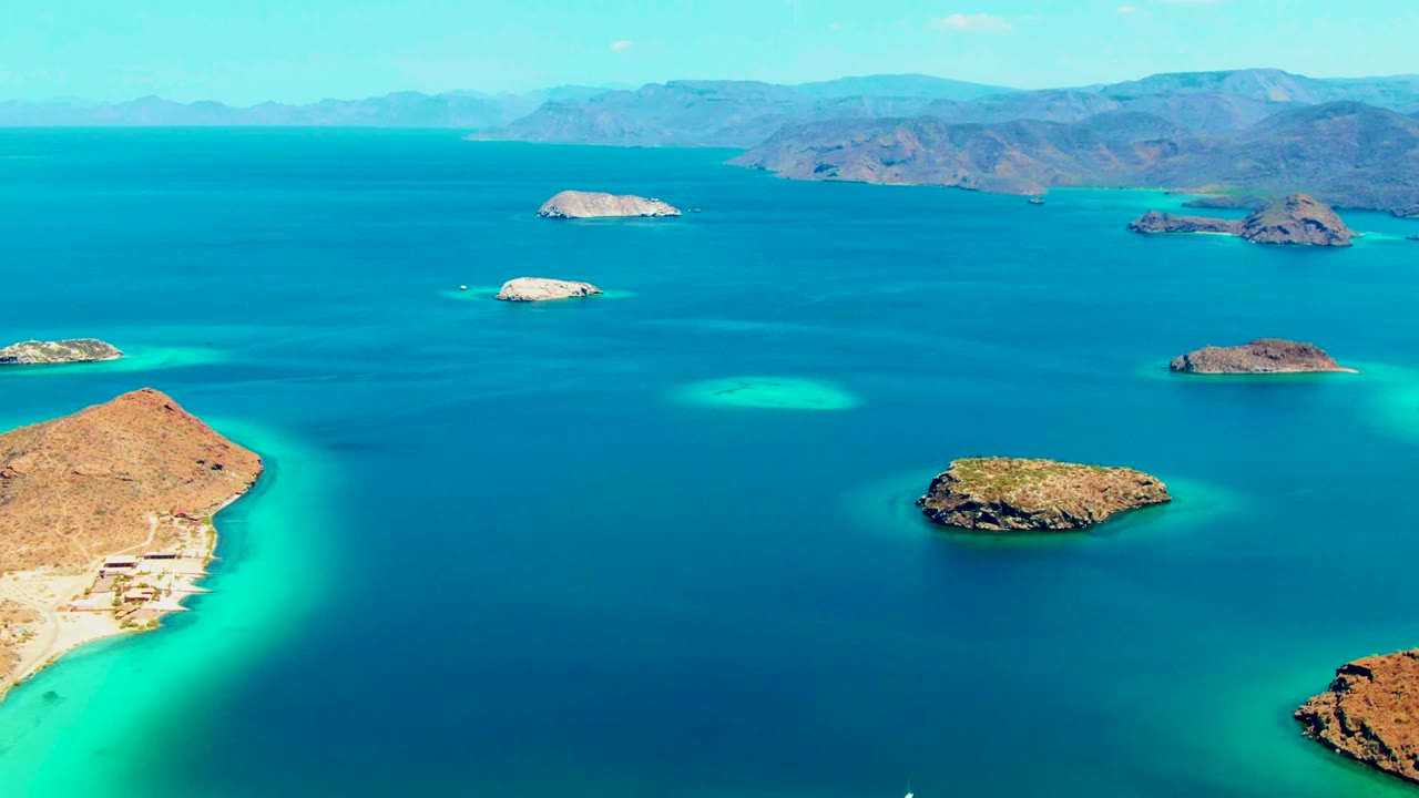 Aerial view at Playa Santispac showing multiple low rocky islands, clear turquoise patches in the water, a mountain ridge beyond the bay, and no visible people.