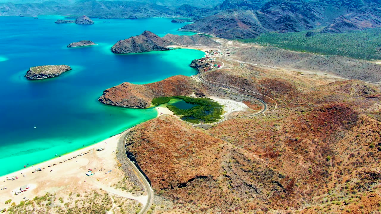 High aerial frame over Playa Santispac showing a sandy beach at lower left, turquoise shallows, rocky islands in the bay, a winding road, dry hills, and no visible people.