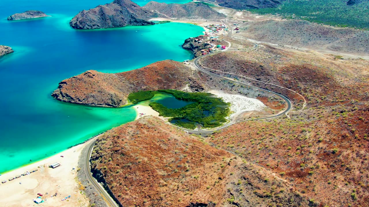 Aerial frame at Playa Santispac, Baja California Sur, showing a sandy beach beside turquoise water, a curving road, dry rocky hills, small vehicles, tiny people, and a green lagoon-like shoreline patch.
