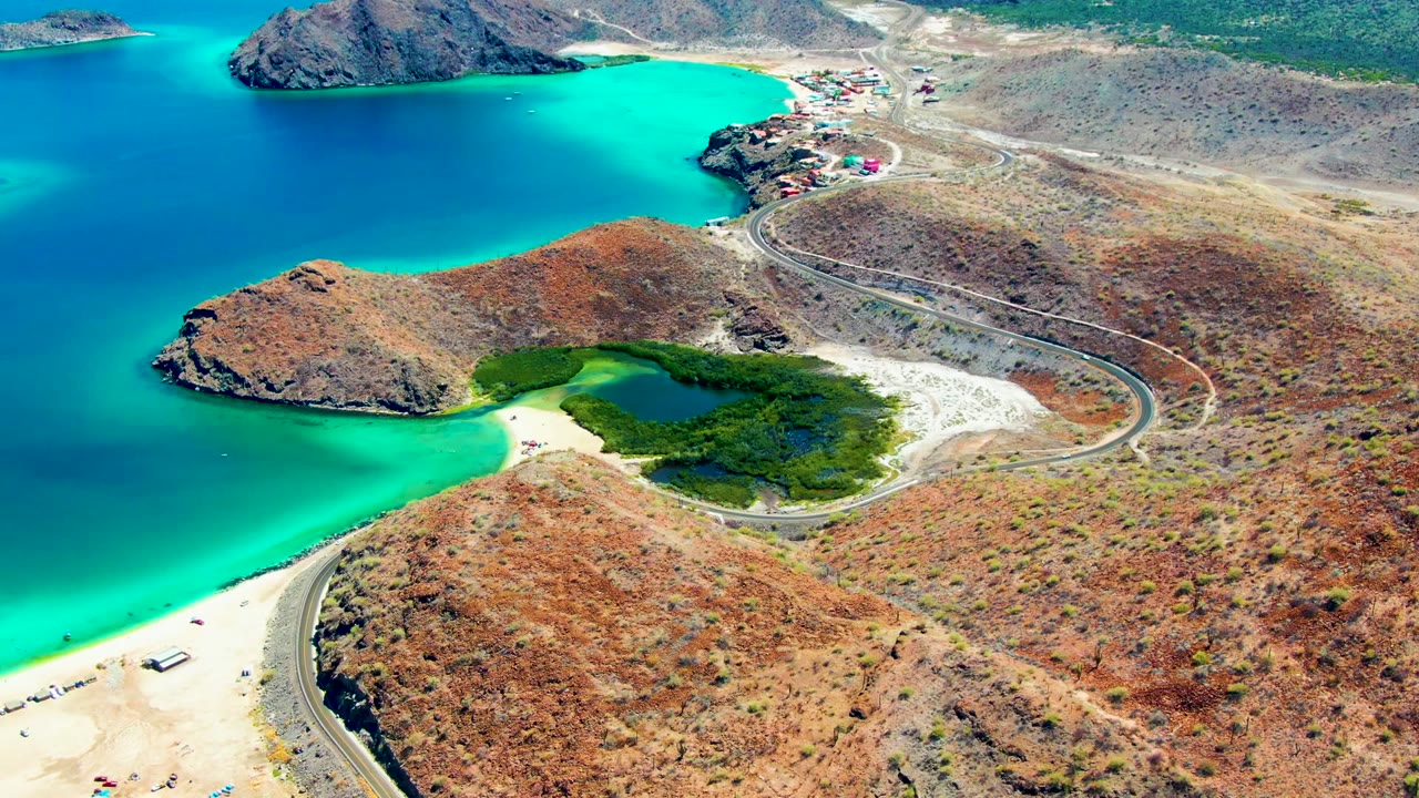 Aerial view of a curving coastal road beside sand, turquoise water, dry hills, vehicles, and a green patch near shore.