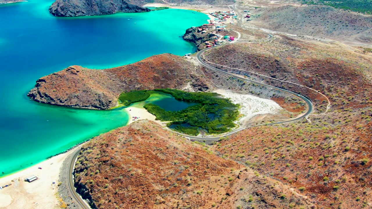 At 0:02, the aerial frame shows more of the road running past dry hills and scattered buildings at Playa Santispac, with turquoise water and a pale sandy beach below.