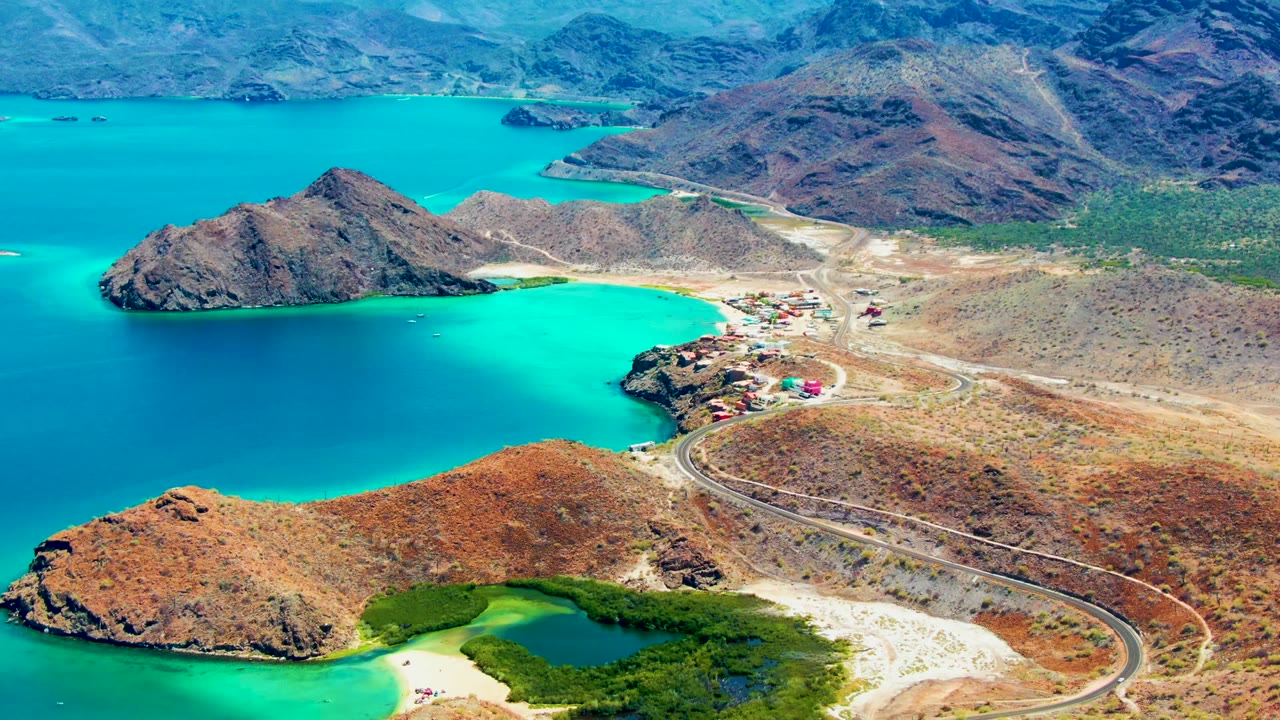 Aerial view over Playa Santispac showing a winding road, rocky dry hills, turquoise water, a small shoreline settlement, distant mountains, and no visible people.