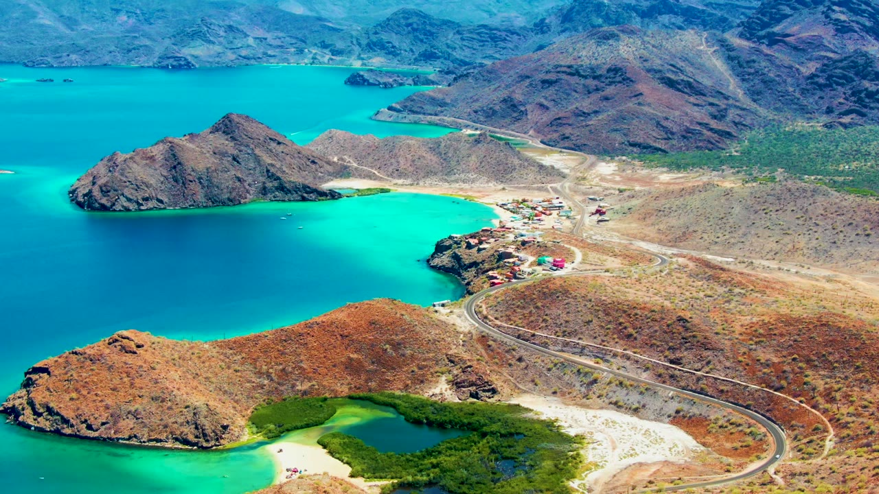 Wide aerial frame of a curving road, settlement, turquoise water, and rocky shoreline.
