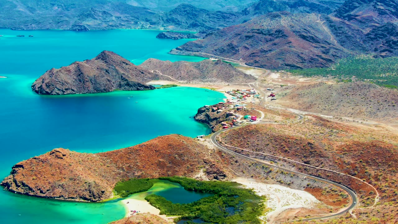 Playa Santispac appears from above with turquoise water wrapping rocky points, a road bending through dry ground, small buildings near shore, and no visible people.
