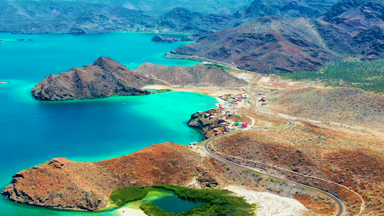 Wide aerial view of a cove with turquoise water, rocky peninsulas, and a shore road.