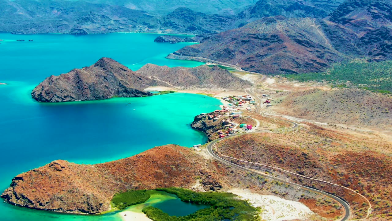 At 0:02, Playa Santispac appears from above with bright blue-green water, dry hills, a shoreline settlement, a curved road, and no visible people.