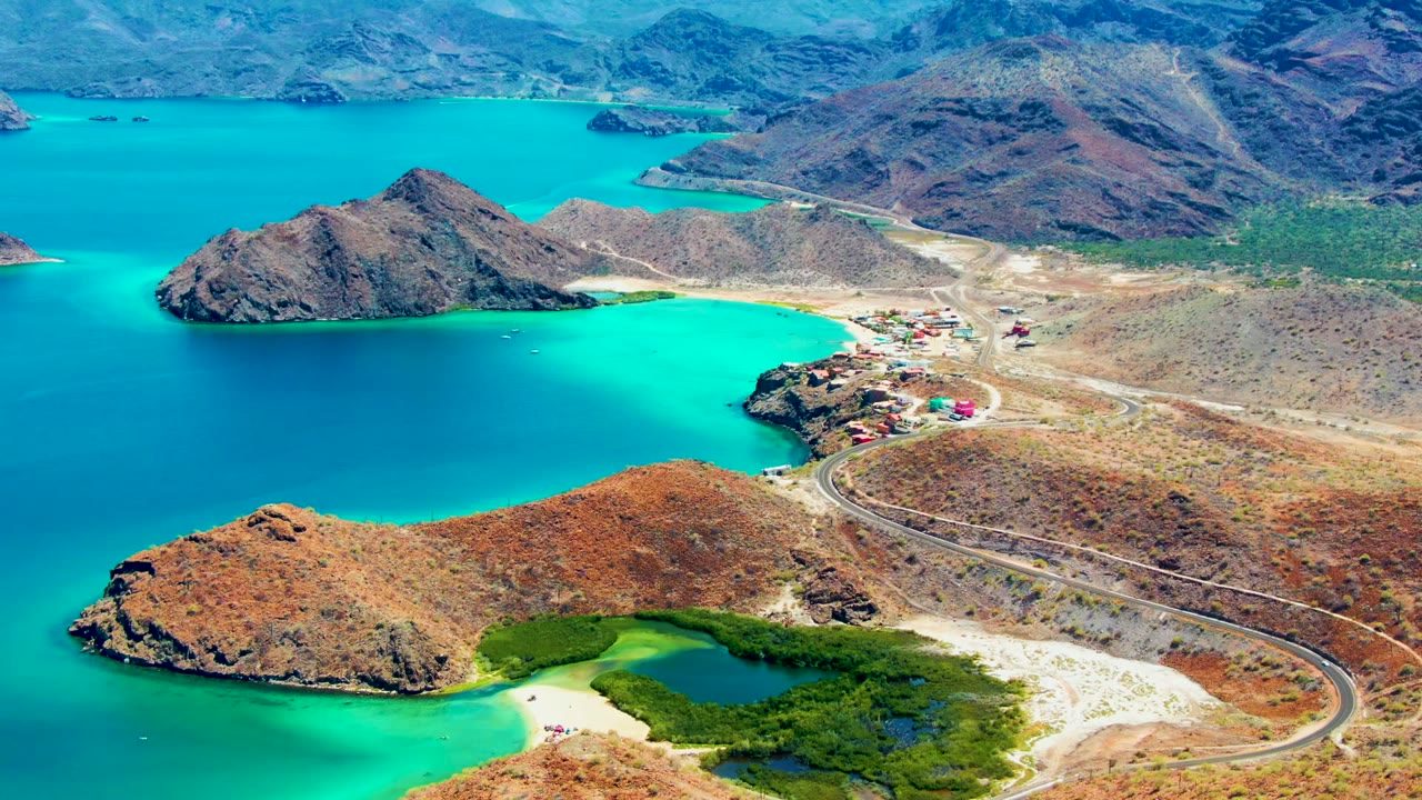 Aerial frame over Playa Santispac, Baja California Sur, showing turquoise water around rocky islands, a curving road, pale beach edges, dry hills, and no visible people.