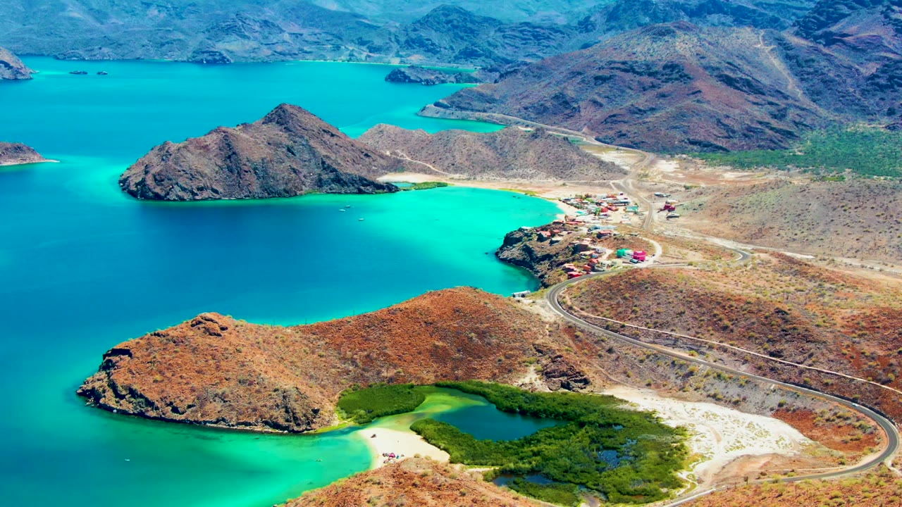Wide aerial frame over Playa Santispac showing a rocky island in turquoise water, the coastal road sweeping past a small settlement, arid hills, distant mountains, and no visible people.