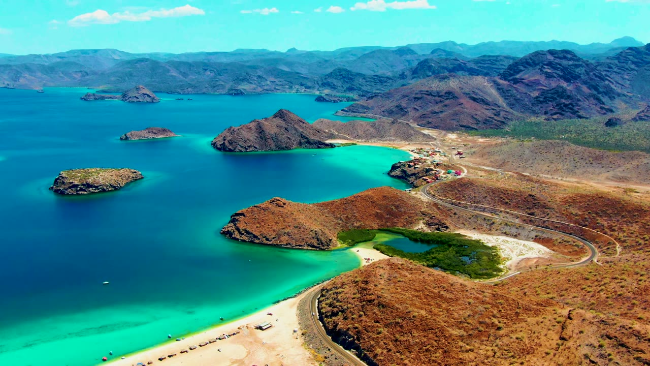 Third aerial frame at Playa Santispac showing green vegetation beside the beach, a tan road curving around dry hills, rocky islands in blue water, distant mountains, and no visible people.