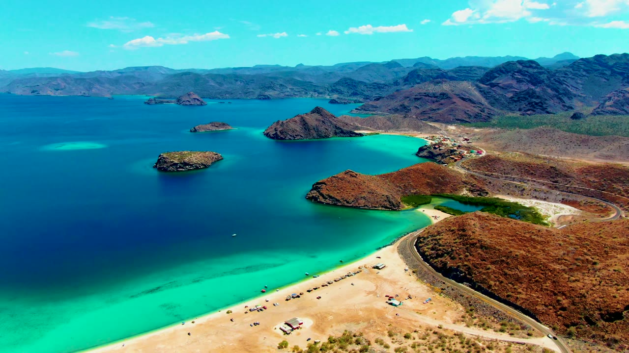 Aerial frame over Playa Santispac showing bright turquoise shallows, deep blue bay water, a sandy beach, rocky islands, distant mountains, and no visible people.