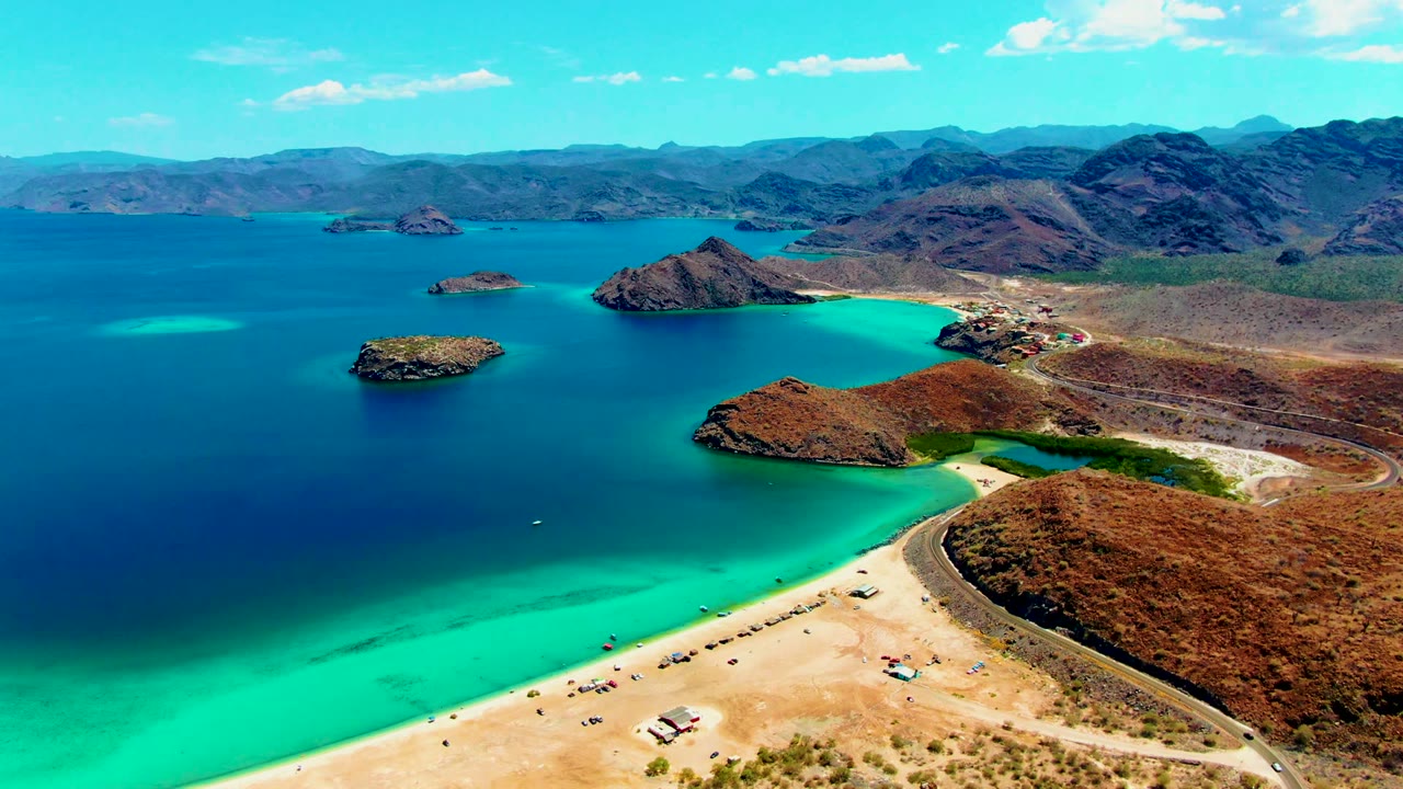 Wide aerial coastline with turquoise shallows, beach road, small boats, and rocky islands.