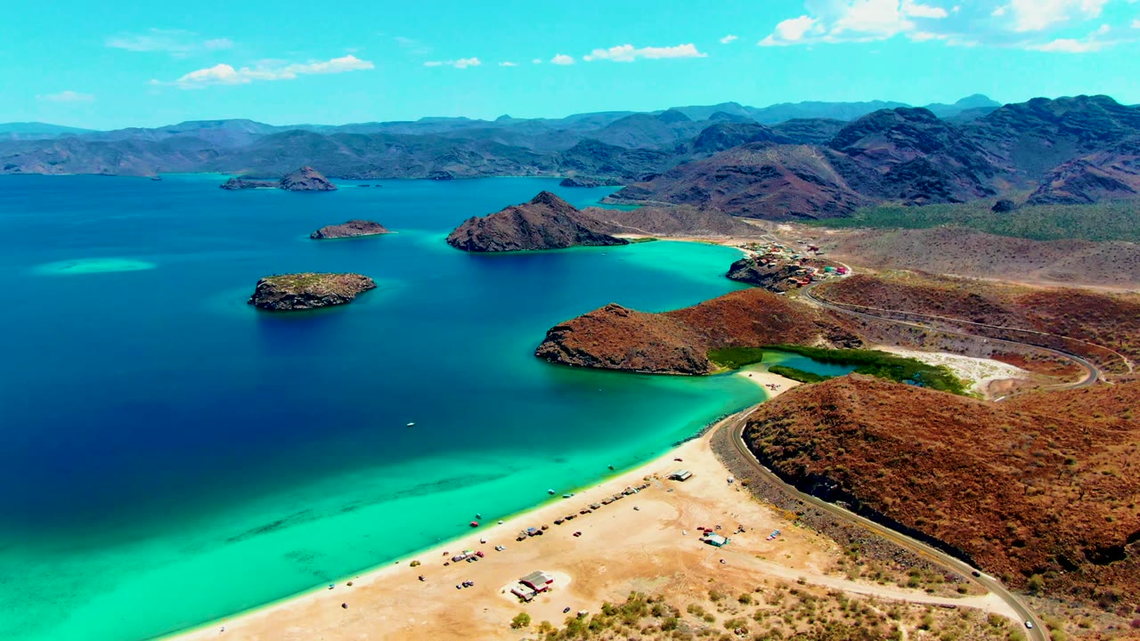 Aerial frame from Playa Santispac showing a pale beach at lower right, a dark road along dry hillside, blue-green bay water, rocky islands, and no visible people.