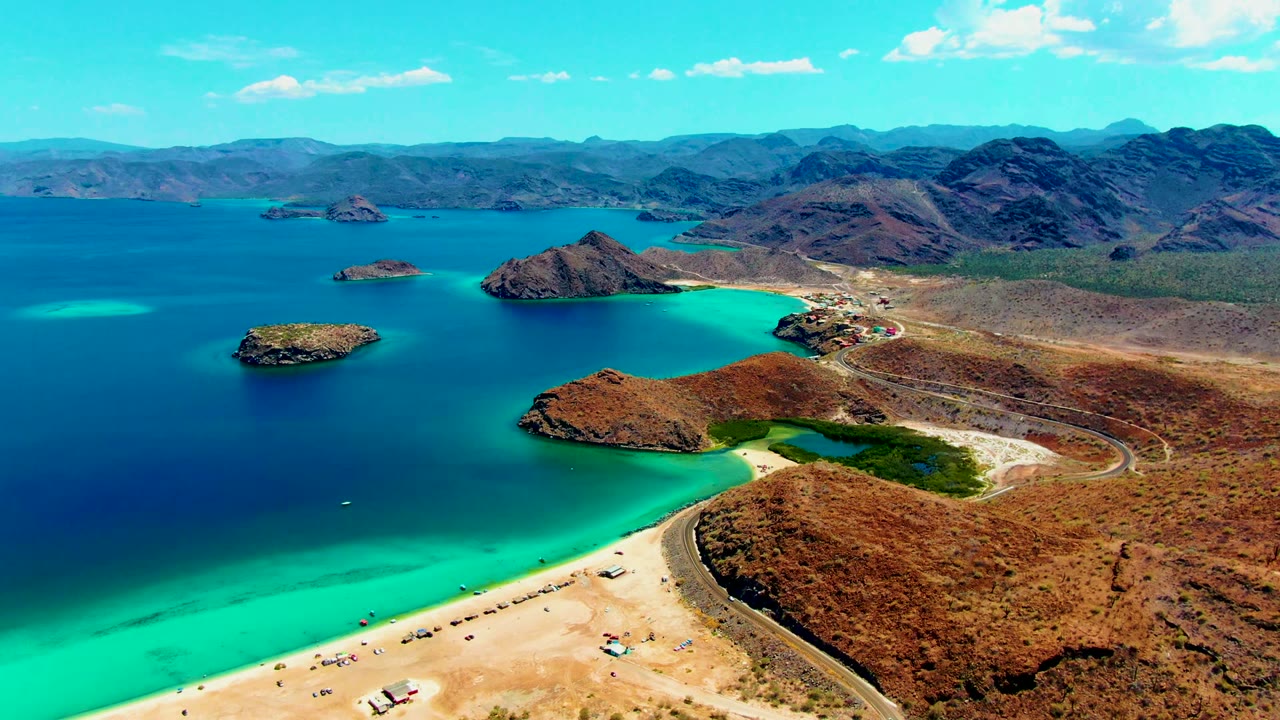 A high aerial frame over Playa Santispac shows turquoise shallows along the sandy beach, darker blue bay water, rocky islands offshore, a curved road on dry terrain, distant mountains, and no visible people.