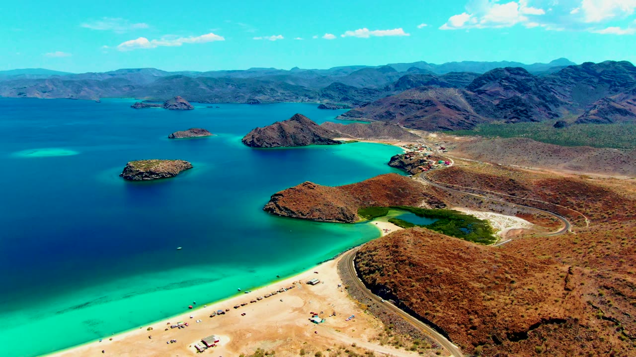 Wide drone view of sandy beach beside turquoise water and rocky islands.