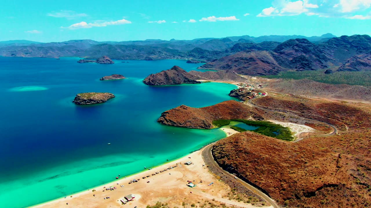 At 0:02, the aerial composition shows Playa Santispac with a curved sandy beach in the lower frame, vehicles near the shore, a road tracing the desert edge, turquoise water, rocky islands, mountains, and no visible people.