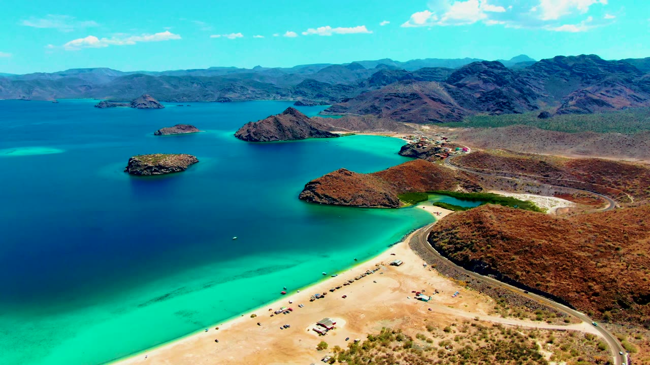 Opening aerial frame over Playa Santispac with a sandy beach and curved road in the foreground, turquoise shallows, deep blue bay, rocky islands, distant mountains, and no visible people.