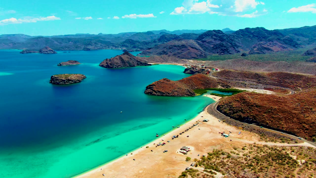 Opening aerial frame of Playa Santispac with a pale sandy beach at lower right, turquoise shallows, dark blue bay water, small offshore islands, a shoreline road, and distant dry mountains.