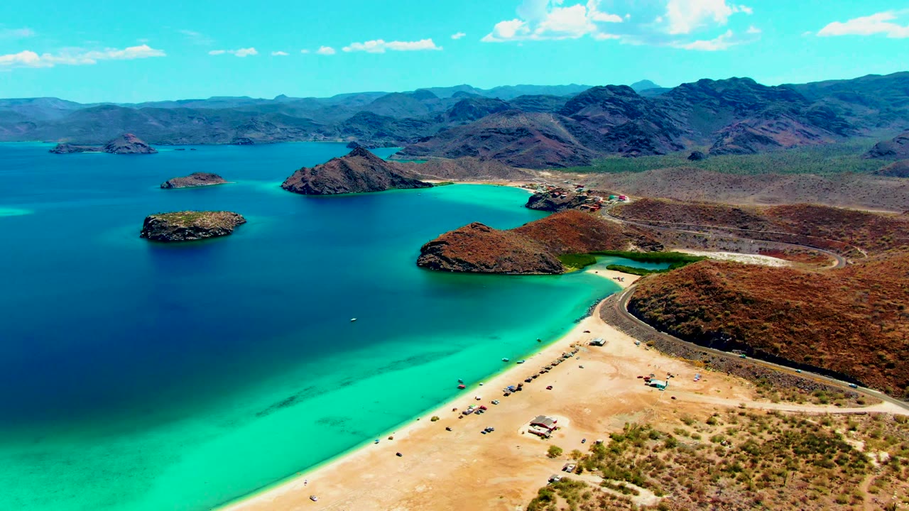 Wide drone view of beachgoers and vehicles beside a turquoise cove.